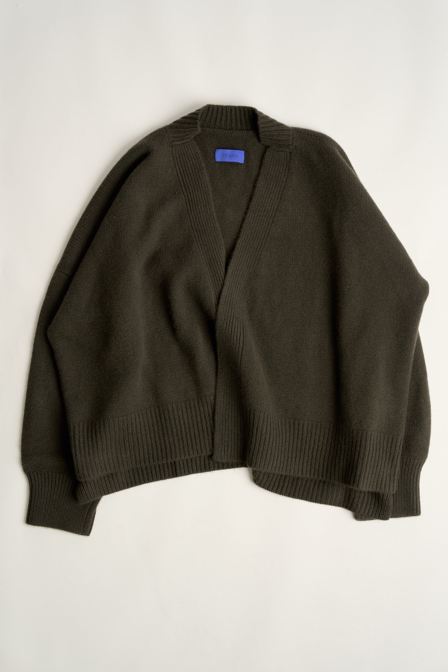 This image features the Aire Cardigan, a luxurious, chunky open cardigan made from pure cashmere. The cardigan is displayed on a model, showcasing its sleek forest color and stylish design elements.