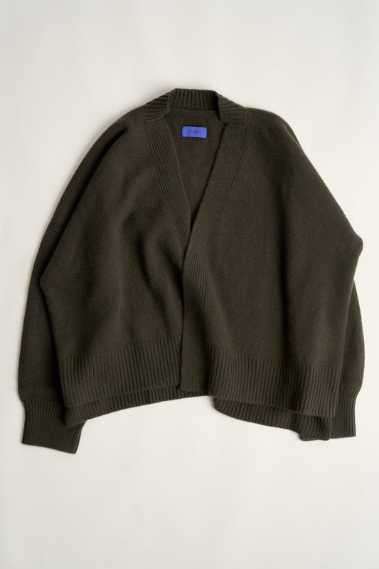 This image features the Aire Cardigan, a luxurious, chunky open cardigan made from pure cashmere. The cardigan is displayed on a model, showcasing its sleek forest color and stylish design elements.