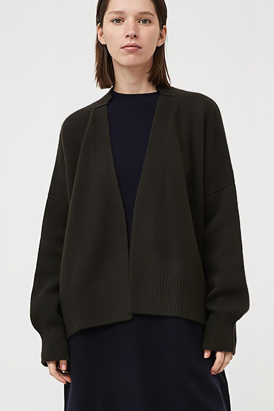 This image features the Aire Cardigan, a luxurious, chunky open cardigan made from pure cashmere. The cardigan is displayed on a model, showcasing its sleek forest color and stylish design elements.