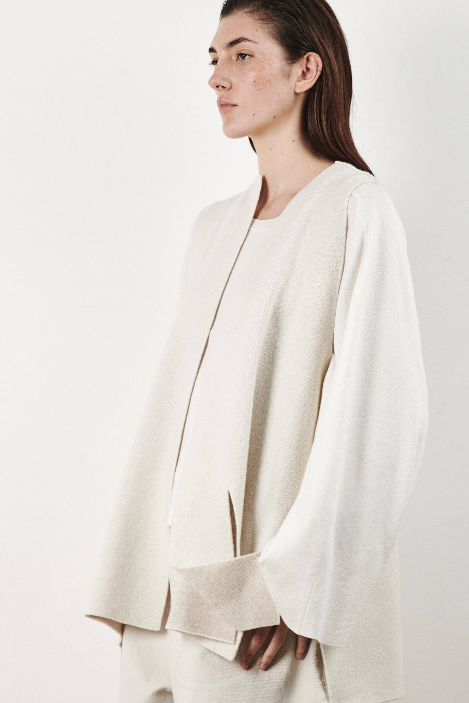 This image features the Ayal Jacket, a stylish sleeveless cashmere jacket displayed on a female model. The jacket is characterized by its asymmetric design, with a distinctive slit on the front left panel and button details on the front hem, adding a touch of uniqueness to its appearance.