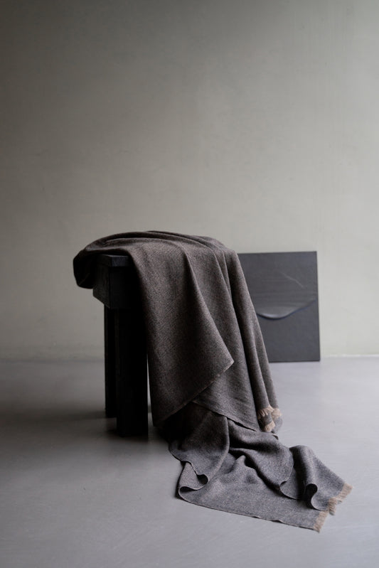 A charcoal Saan throw with a herringbone design is draped over the back of a chair, with the excess hanging elegantly to the side. The throw appears soft and is detailed with a subtle pattern and short fringes.