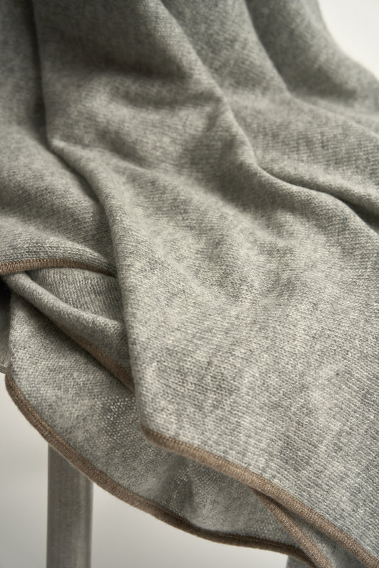 A soft grey throw with a fine contrast color edge draped over a chair, against a white background.