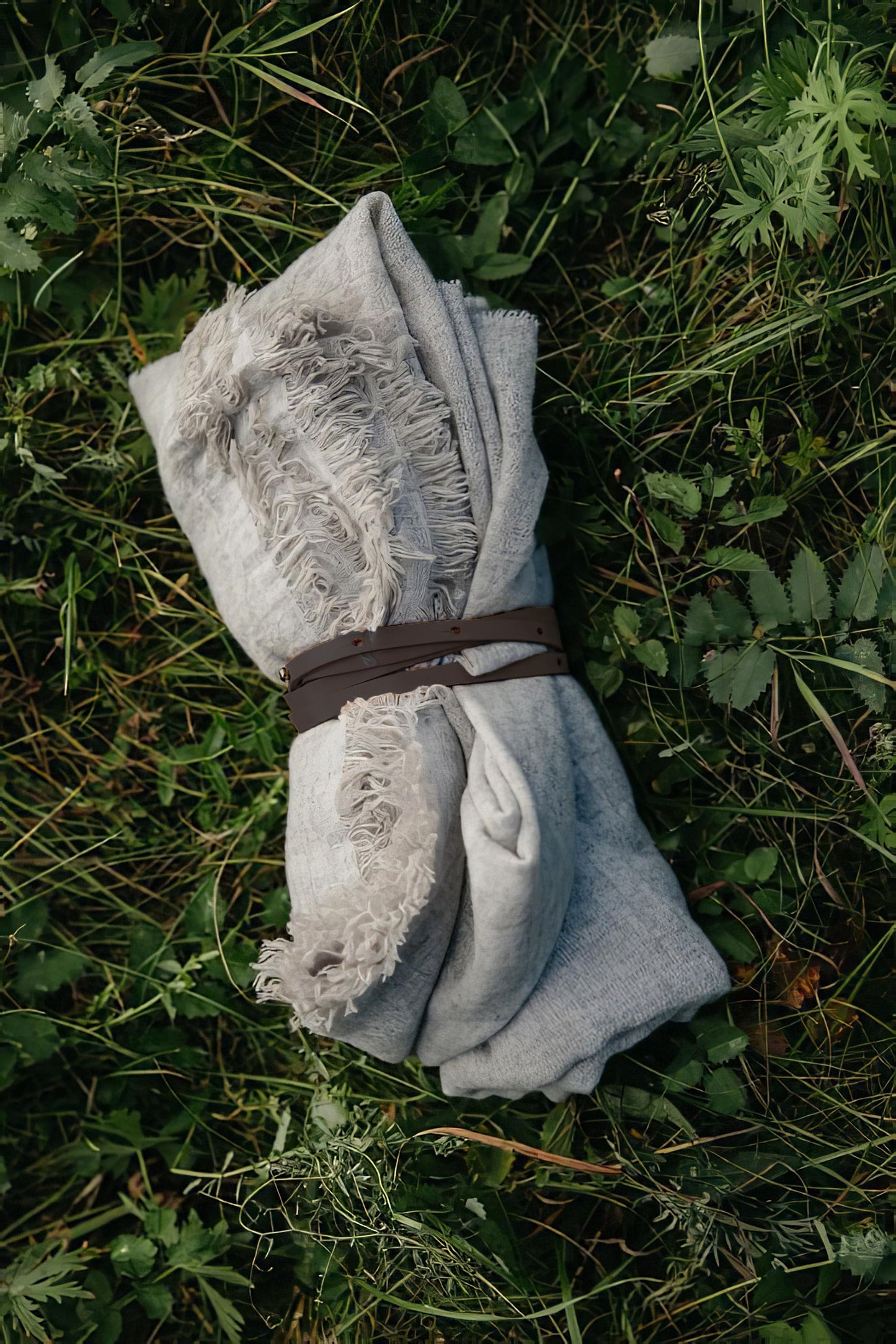 A gray, finely woven travel throw with a fringed edge, laid out on a grassy field with daisies around it, secured with a brown belt.