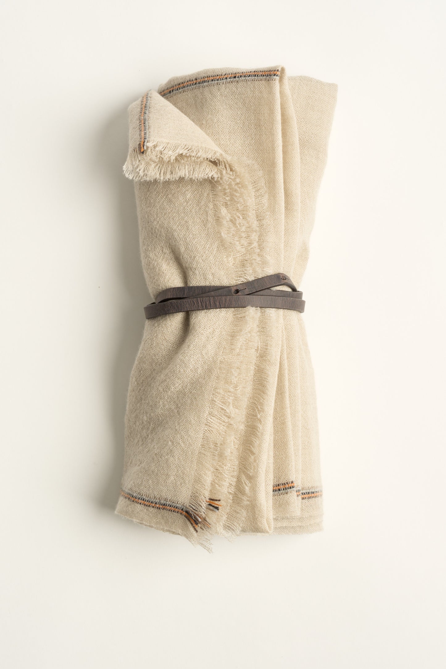 A rolled-up beige cashmere travel throw with visible fringes, secured with a brown leather belt.