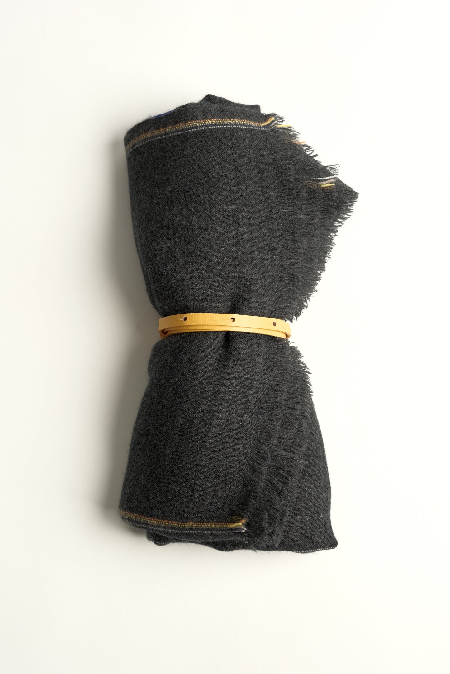 A rolled-up charcoal cashmere travel throw with visible fringes, secured with a gold coloured belt.