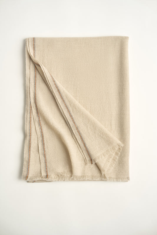 A folded beige-colored cashmere throw with visible contrast stripes and fringes on a light background.
