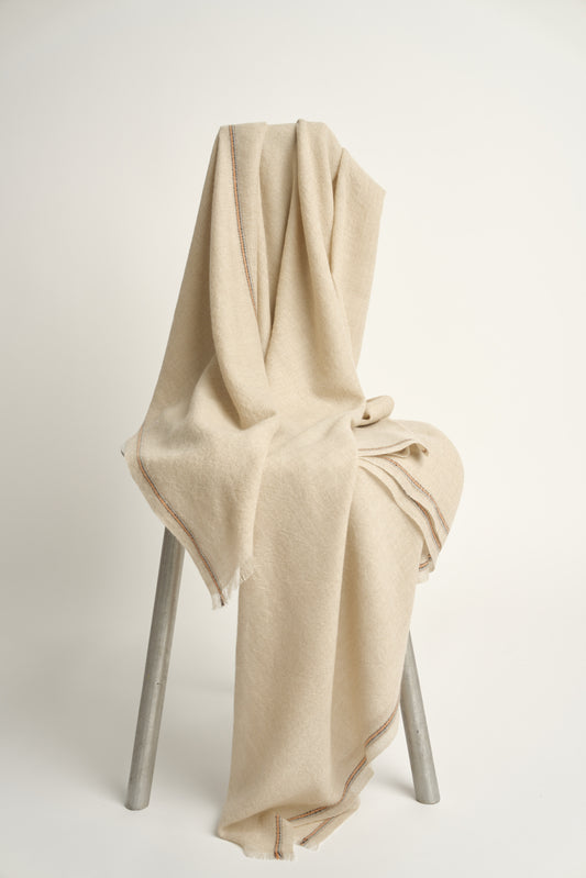 A beige-colored cashmere throw with visible contrast stripes and fringes, draped over a chair on a light background.