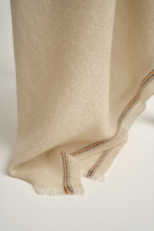 A beige-colored cashmere throw with visible contrast stripes and fringes, draped over a chair on a light background.