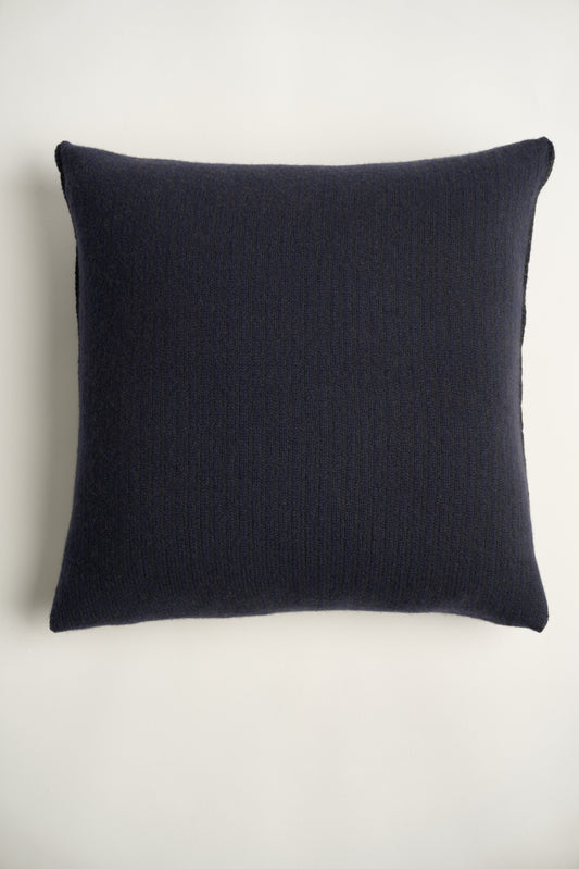 This image displays the Letra Cushion Cover, characterized by its knitted two-tone float knit design. The cushion cover features a textured, cozy appearance in a dark color, which comes across as modern and stylish.