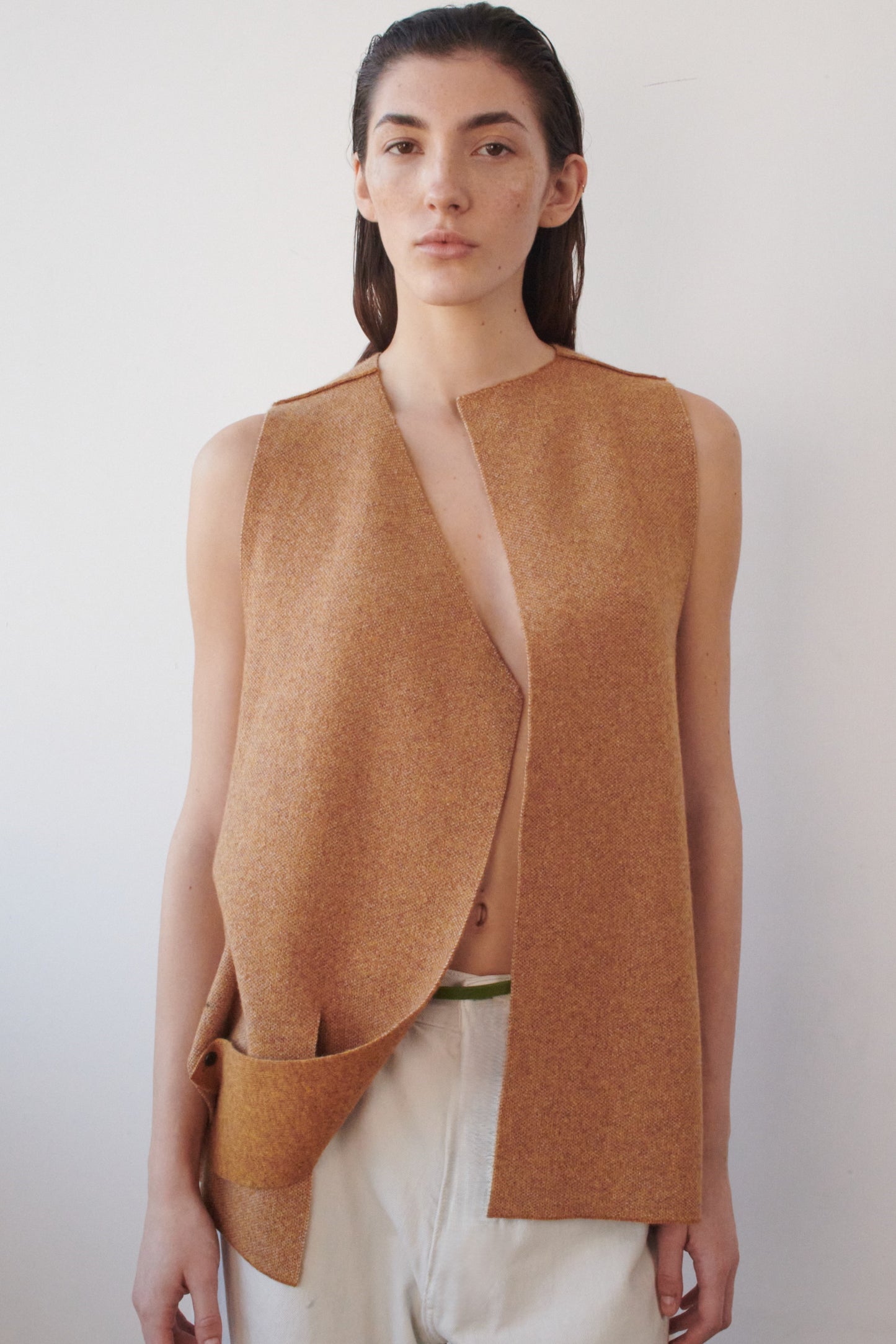 This image features the Ayal Jacket, a stylish sleeveless cashmere jacket displayed on a female model. The jacket is characterized by its asymmetric design, with a distinctive slit on the front left panel and button details on the front hem, adding a touch of uniqueness to its appearance.