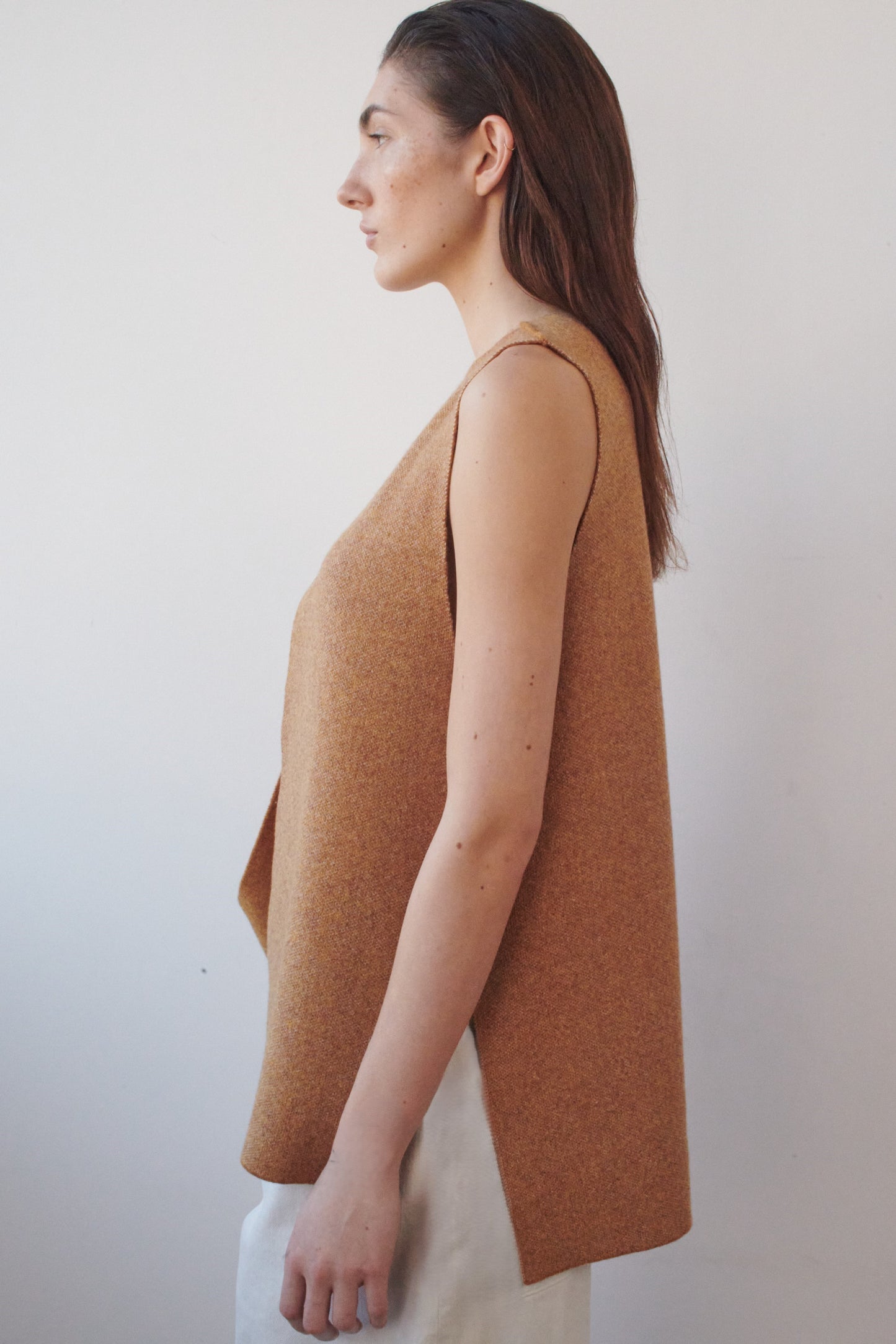 This image features the Ayal Jacket, a stylish sleeveless cashmere jacket displayed on a female model. The jacket is characterized by its asymmetric design, with a distinctive slit on the front left panel and button details on the front hem, adding a touch of uniqueness to its appearance.