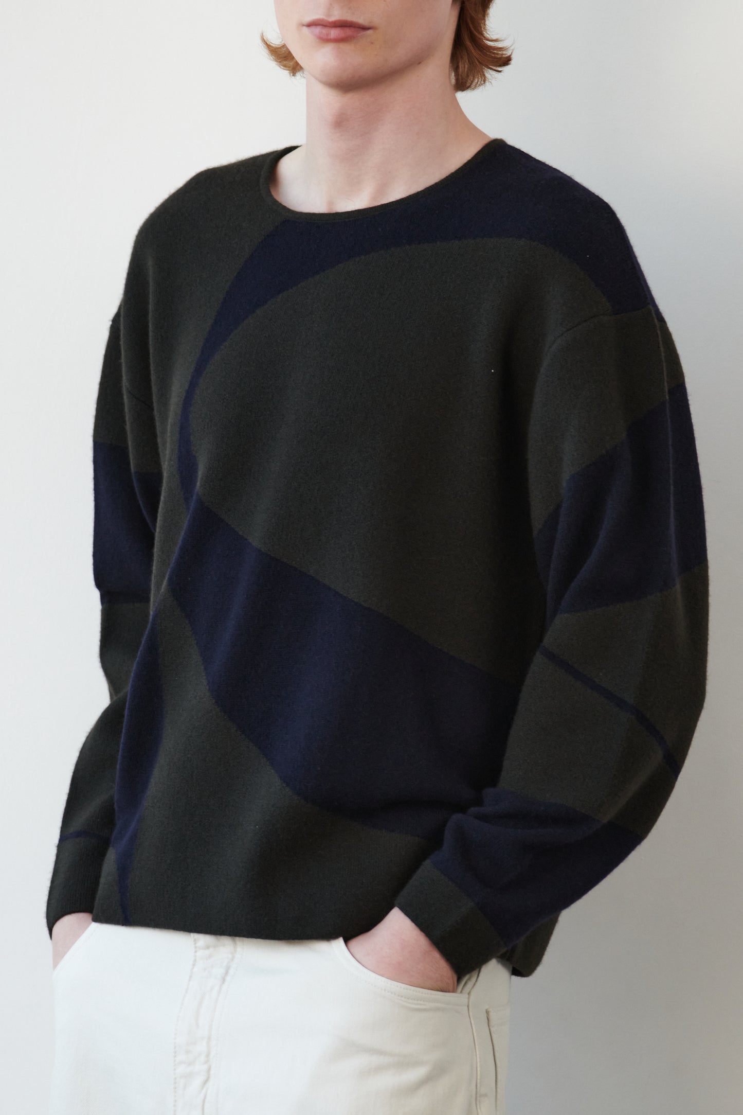 This image features the Arouc Sweater, a stylish and cozy garment that embodies a relaxed, everyday aesthetic. The sweater displays a boxy silhouette, crafted in a knitted jacquard print that forms an abstract, organic pattern in shades of green and navy blue.