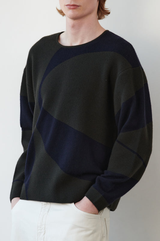 This image features the Arouc Sweater, a stylish and cozy garment that embodies a relaxed, everyday aesthetic. The sweater displays a boxy silhouette, crafted in a knitted jacquard print that forms an abstract, organic pattern in shades of green and navy blue.
