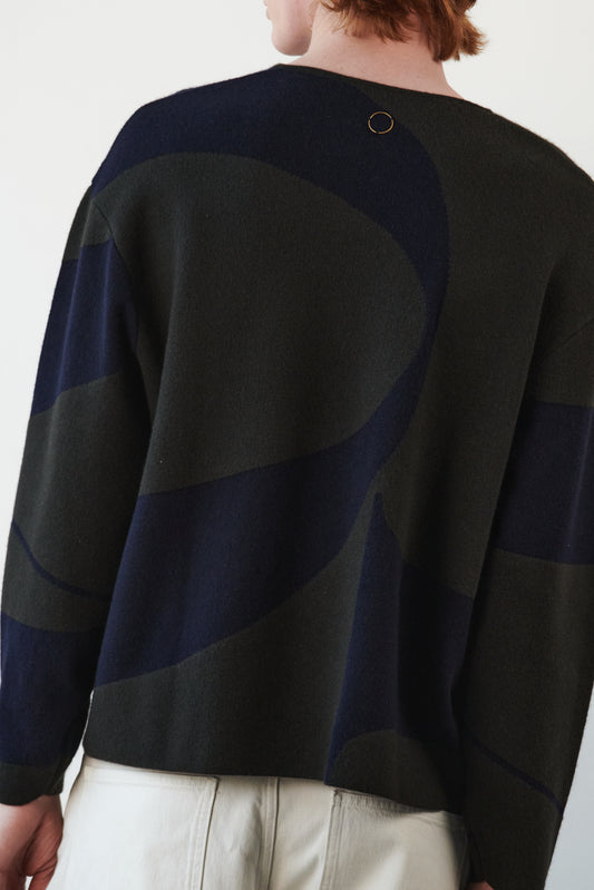 This image features the Arouc Sweater, a stylish and cozy garment that embodies a relaxed, everyday aesthetic. The sweater displays a boxy silhouette, crafted in a knitted jacquard print that forms an abstract, organic pattern in shades of green and navy blue.