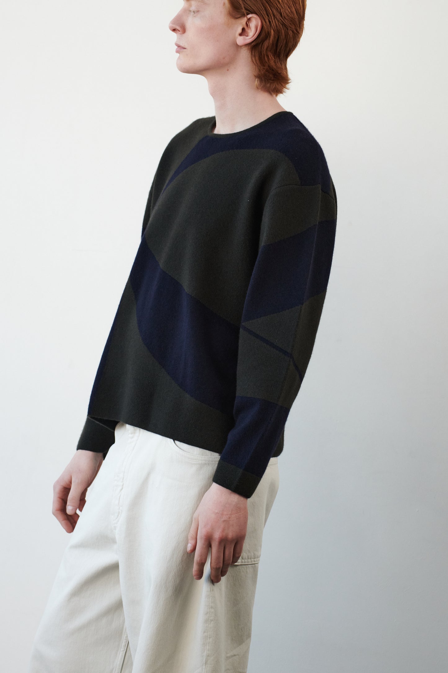 This image features the Arouc Sweater, a stylish and cozy garment that embodies a relaxed, everyday aesthetic. The sweater displays a boxy silhouette, crafted in a knitted jacquard print that forms an abstract, organic pattern in shades of green and navy blue.