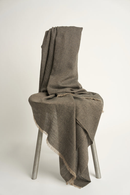 A charcoal and taupe Saan throw with a herringbone design is draped over the back of a chair, with the excess hanging elegantly to the side. The throw appears soft and is detailed with a subtle pattern and short fringes.