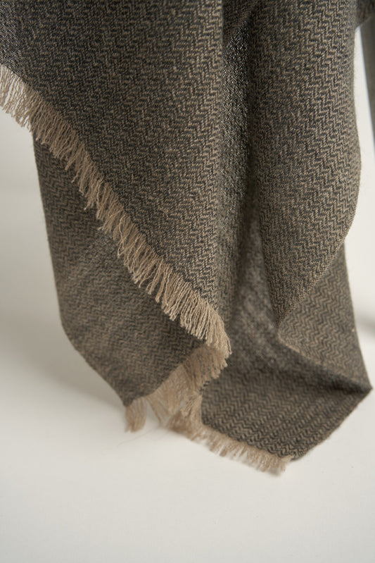 A charcoal and taupe Saan throw with a herringbone design is draped over the back of a chair, with the excess hanging elegantly to the side. The throw appears soft and is detailed with a subtle pattern and short fringes.
