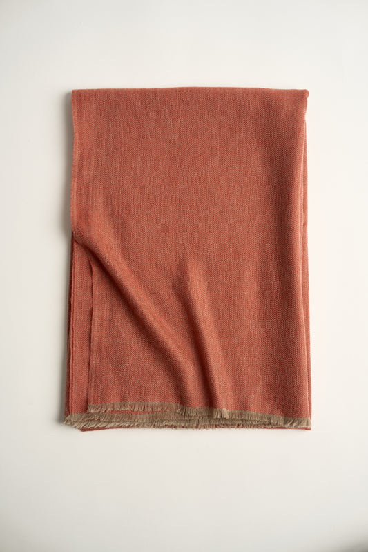 A coral and taupe Saan throw with a herringbone design is draped over the back of a chair, with the excess hanging elegantly to the side. The throw appears soft and is detailed with a subtle pattern and short fringes.