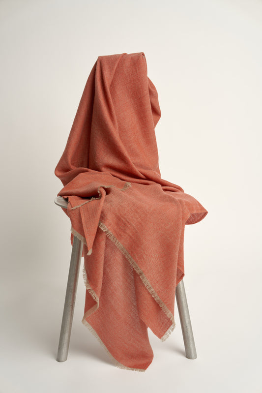 A coral and taupe Saan throw with a herringbone design is draped over the back of a chair, with the excess hanging elegantly to the side. The throw appears soft and is detailed with a subtle pattern and short fringes.
