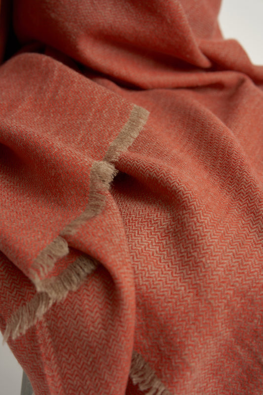 A coral and taupe Saan throw with a herringbone design is draped over the back of a chair, with the excess hanging elegantly to the side. The throw appears soft and is detailed with a subtle pattern and short fringes.