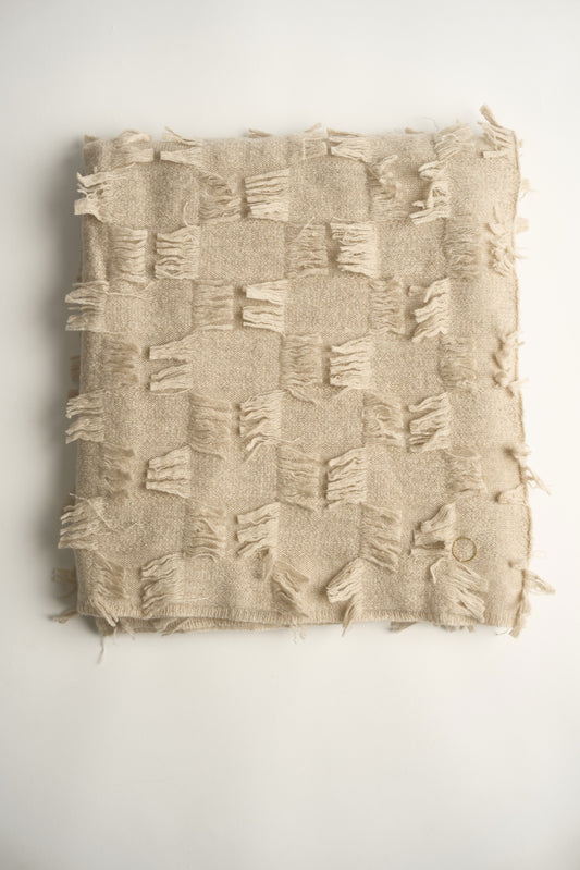 This image displays the Seren Throw, a luxurious cashmere knitted throw featuring a distinctive hand-cut fringe detail. The throw is presented in a bright, warm beige color, enhancing its elegant and sophisticated appearance.