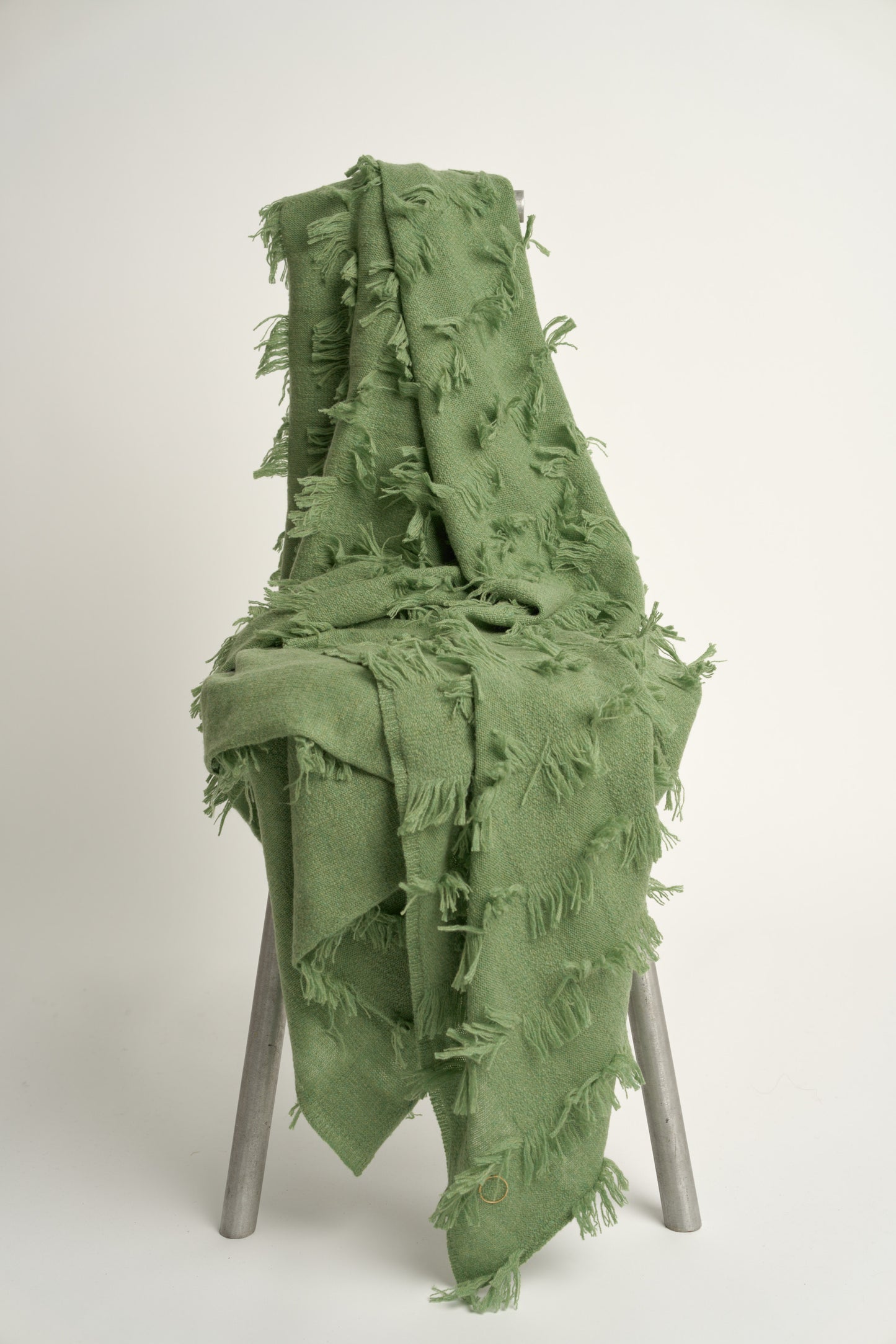 This image displays the Seren Throw, a luxurious cashmere knitted throw featuring a distinctive hand-cut fringe detail. The throw is presented in a natural, embracing green color, enhancing its elegant and sophisticated appearance.