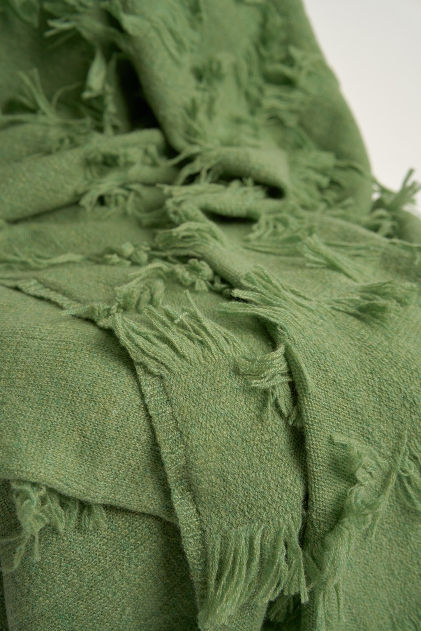 This image displays the Seren Throw, a luxurious cashmere knitted throw featuring a distinctive hand-cut fringe detail. The throw is presented in a natural, embracing green color, enhancing its elegant and sophisticated appearance.