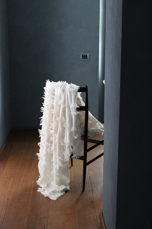 This image displays the Seren Throw, a luxurious cashmere knitting throw featuring a distinctive hand-cut fringe detail. The throw is presented in a light, bright ivory color, enhancing its elegant and sophisticated appearance.