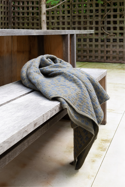 This image features the "stone throw in moss & slate," a luxurious and meticulously crafted cashmere throw. The throw displays an elegant pattern that resembles the texture of slate floors, complemented by bands of yarn in varying lengths that add a subtle yet dynamic visual appeal.