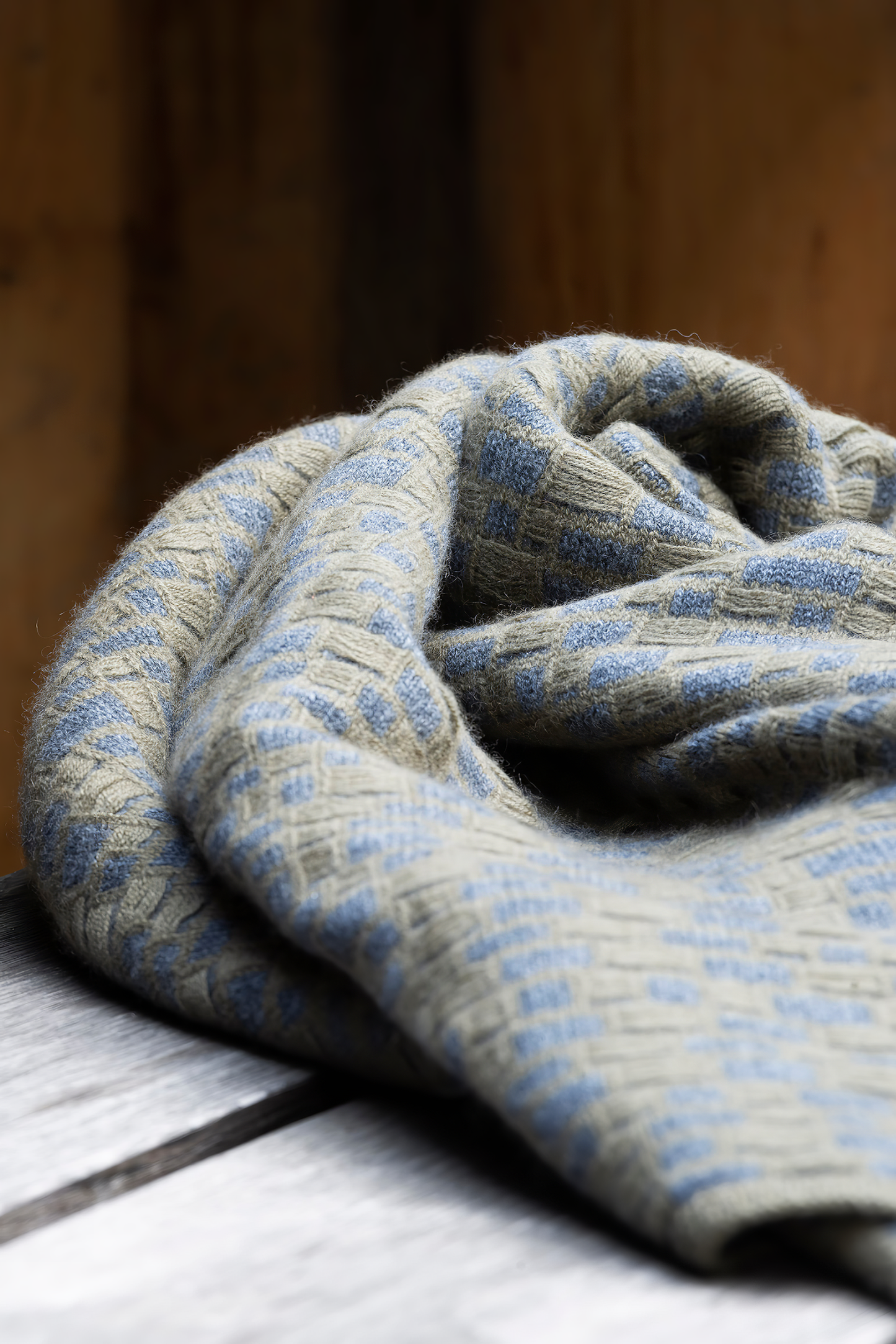 This image features the "stone throw in moss & slate," a luxurious and meticulously crafted cashmere throw. The throw displays an elegant pattern that resembles the texture of slate floors, complemented by bands of yarn in varying lengths that add a subtle yet dynamic visual appeal.