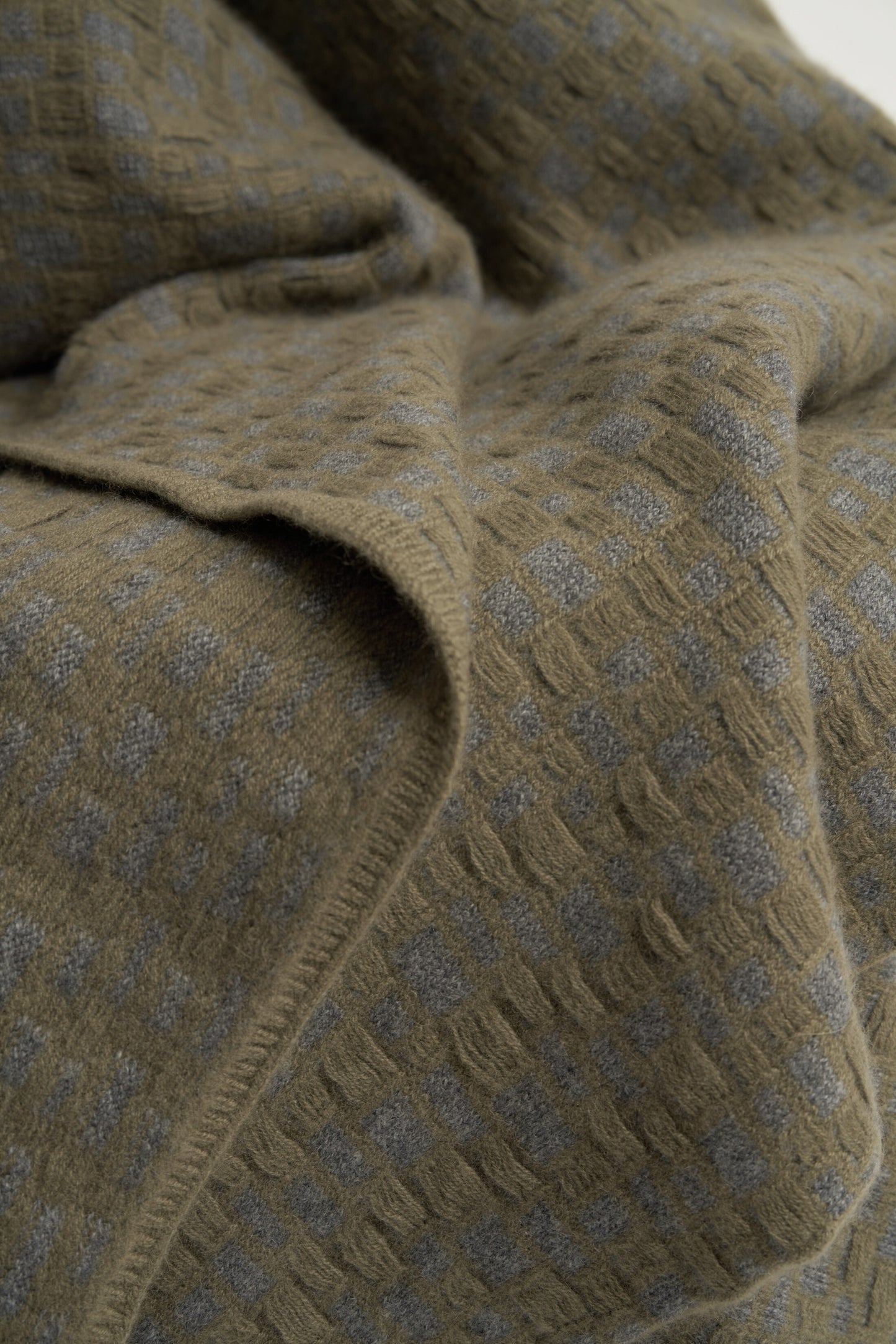 This image features the "stone throw in moss & slate," a luxurious and meticulously crafted cashmere throw. The throw displays an elegant pattern that resembles the texture of slate floors, complemented by bands of yarn in varying lengths that add a subtle yet dynamic visual appeal.