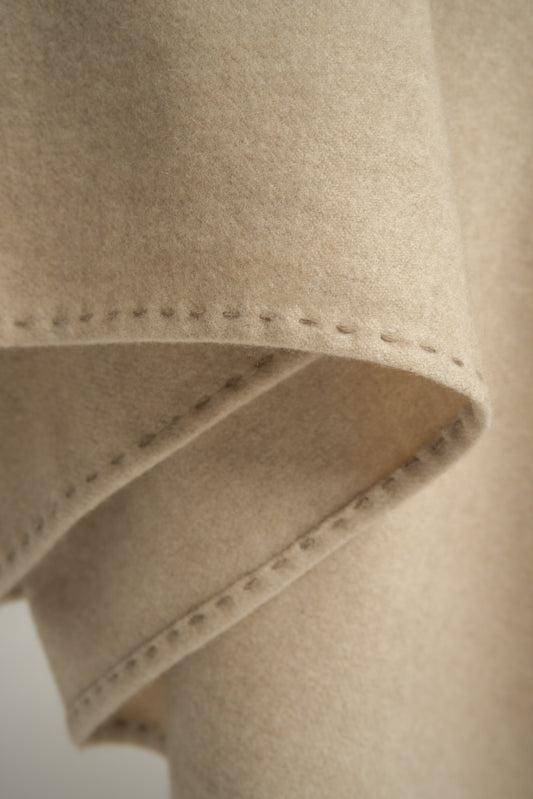 A beige Suono throw with hand-stitched edges, folded over a chair and displayed on a white background.